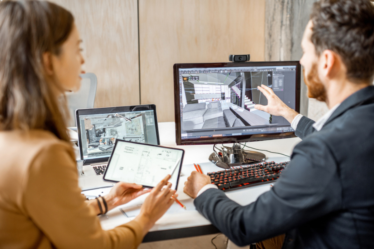 Zdjęcie przedstawia kobietę i męższczyzny przy biurku pełnym komputerów. Tłumaczą coś sobie